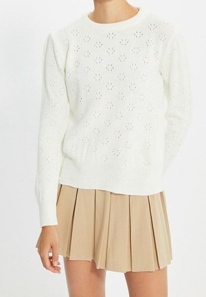 White knit sweater with small eyelet patterns paired with a beige pleated mini skirt on a standing person.
