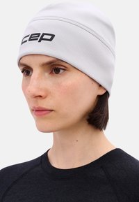 Jong persoon draagt een witte beanie met het "cep" logo, donker kort haar en een zwart shirt met ronde hals, kijkt iets opzij.