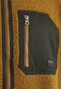 Bruine, gestructureerde fleecejack met een zwarte fabric patch pocket voorzien van een verticale ritssluiting en een klein merkje.