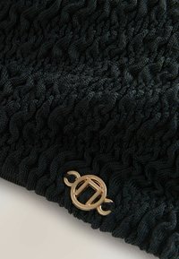 Zwart gebreide stof met een structuur en golvend patroon. Heeft een gouden ronde accent aan de zijkant bevestigd. Zacht, rekbaar materiaal.