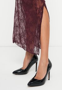 Sieviete, kas valkā melnas lakotas ādas augstpapēžu kurpes un burgundijas krāsas mežģīņu svārkus ar augstu šķēlumu, stāv uz baltas grīdas pret baltu fona.