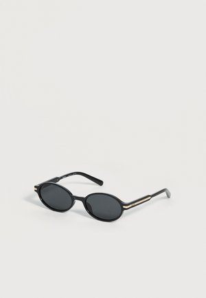Lunettes de soleil ovales noires avec des fines montures et des verres foncés reposant sur une surface gris clair.
