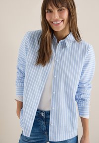 Jeune femme souriante portant une chemise à rayures verticales bleu clair et blanc sur un haut blanc et un jean bleu.