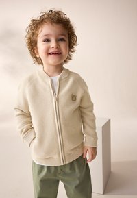 Cream zip-up jacket with textured fabric, featuring side pockets and a small bear logo on the chest, paired with olive green pants.