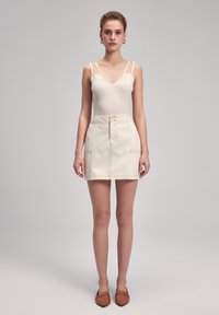 Cremefarbene Jeans-Minirock mit vorderem Knopfverschluss, zwei Fronttaschen und kontrastierender Naht. Das Modell trägt ein geripptes Tanktop.