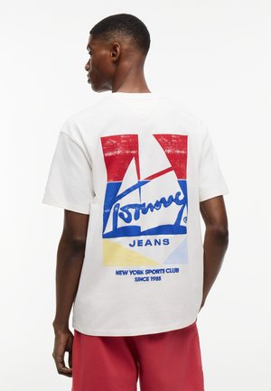 Hombre de espaldas lleva una camiseta blanca con un gráfico y texto en rojo, azul y amarillo que dice "Nieuw York Sports Club Desde 1985" y pantalones cortos rojos.