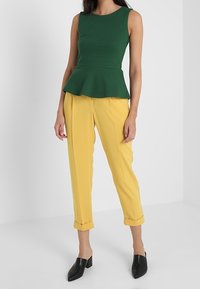 Zelený top s peplum stylem z texturované látky, zkombinovaný se žlutými úzkými kalhotami. Černé muly s špičatou špičkou. Jednoduchý, moderní design.