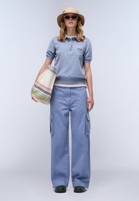 Lichtblauwe polo met korte mouwen, geborduurd logo, gecombineerd met losse lichtblauwe cargo broeken. Beige zonnenhoed en gestreepte tote bag.