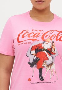 Rosa grafisk t-skjorte med julefe, Santa Claus i en stol, som holder en Coca-Cola-flaske, med en reinsdyr ved siden av. Tydelige røde trykkdetaljer.