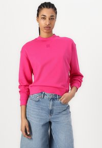 Sudadera rosa con cuello redondo y puños acanalados, que presenta un sutil logo bordado. Combinada con unos vaqueros de denim de pierna ancha en azul claro.