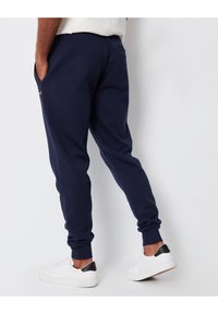 Marineblå joggerbukser med en blød tekstur, elastiske mansjetter og sidelommer. Ryggen har en lille logodetalje. Båret med hvide sneakers.