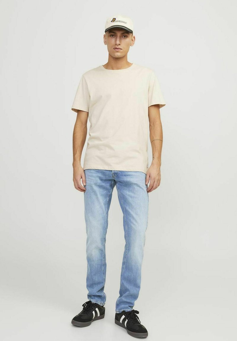 Beige Baumwoll-T-Shirt kombiniert mit hellblauen Denim-Jeans. Schwarze Sneakers mit weißen Streifen und eine beigefarbene Mütze mit Logo-Detail.