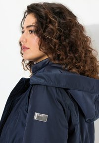 Giacca blu navy con colletto alto, cappuccio ampio e tessuto strutturato. Presenta un'etichetta argentata sulla manica e tiranti regolabili.