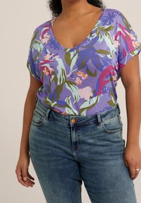 Fialový krátký top s květinovým vzorem a výstřihem do V, zdobený zelenými listy a růžovými květy. Kombinován s modrými džínami s vysokým pasem.