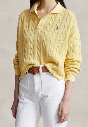 Personne portant un pull jaune en maille torsadée avec col et un pantalon blanc taille haute avec une ceinture marron sur un fond uni.