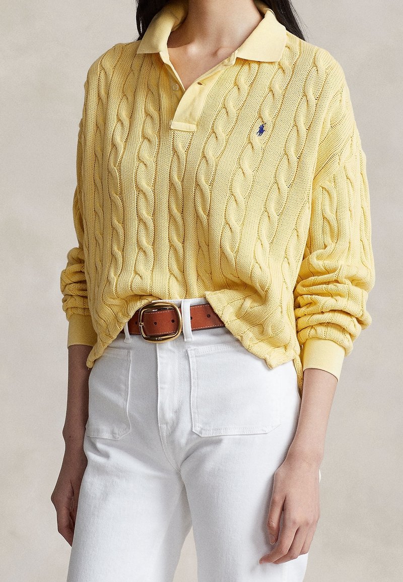 Personne portant un pull jaune en maille torsadée avec col et un pantalon blanc taille haute avec une ceinture marron sur un fond uni.