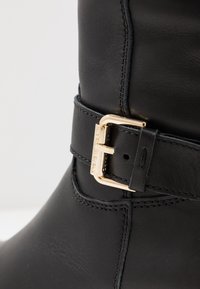 Botín de cuero negro con un acento de hebilla dorada, textura suave y costuras reforzadas. El diseño incluye una correa alrededor del tobillo.