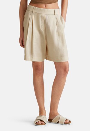 Shorts en lin beige avec plis et poches latérales. L'ourlet se termine juste au-dessus du genou. Associés à des sandales souples à brides croisées.