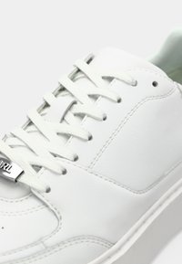 Hvit skinnsneaker med flate lisser, metalldetalj med logo og en strukturert gummisåle. Minimalistisk design med en glatt finish.