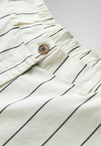 Close-up van crèmekleurige broek met zwarte verticale strepen, voorzien van een elastische tailleband en een bruine knoop aan de voorste gulp.
