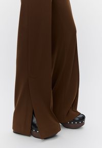 Pantalon large marron à jambes larges avec des fentes latérales et des chaussures à plateforme noire épaisses ornées de clous argentés. Texture de tissu lisse et lignes épurées.