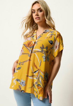 Kvinde iført løs gul blomsterbluse med korte ærmer, kombineret med lyseblå jeans, mod en ensfarvet lys baggrund.