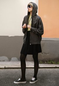 Šedá mikina na zip s kapucí, černá plisovaná sukně, průsvitné černé punčochy a černé tenisky s bílými tkaničkami. Velké sluneční brýle dotvářejí celý vzhled.