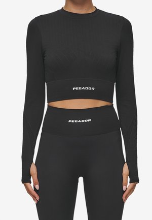 Woman wearing black long-sleeve crop top and high-waist leggings, both with white "PEGADOR" logo, standing with hands relaxed at sides.