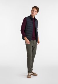 Navy Puffer-Weste über einem burgunderfarbenen Pullover, kombiniert mit olivgrünen Hosen und braunen Sneakers mit hellen Schnürsenkeln. Glatte Textur, Reißverschluss.