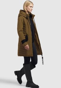 Parka marrone imbottita con cappuccio, dotata di tasche laterali e polsini a coste. Abbinata a leggings neri e stivali alti fino al ginocchio con suola spessa.