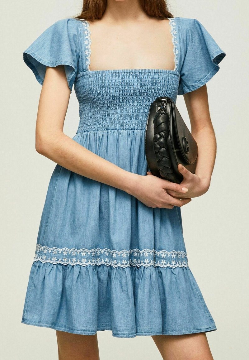 Femme portant une robe bleue smockée à manches courtes avec bordure en dentelle blanche, tenant une pochette en cuir noir tressé au niveau de la taille.