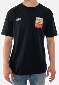 Black cotton t-shirt with short sleeves featuring a colorful printed patch and logo on the left chest area.