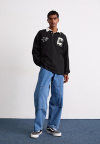 Svart långärmad polo med vit krage, som har en stor 'C' logo och textdesign; blå baggyjeans och grå sneakers.