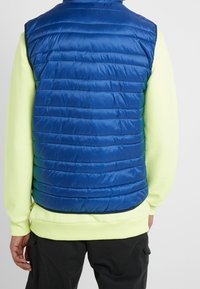 Niebieska puchowa kamizelka z poziomymi przeszyciami, wykonana z błyszczącego, syntetycznego materiału. Noszona na jaskrawożółtej bluzce z długim rękawem.