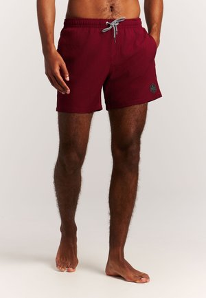 Man draagt bordeaux zwemshorts met zwart-wit trekkoord, staat blootsvoets tegen een effen lichte achtergrond.