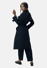 Abrigo negro de corte trench con cintura ceñida, que presenta un amplio cuello y mangas largas. Combinado con pantalones negros recortados y zapatos negros planos.