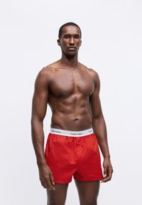 Raudoni šortai boxeriai, pagaminti iš lygaus audinio, su balta elastinga juosta, ant kurios yra "Calvin Klein" logotipas. Modelis stovi su neutralia išraiška.