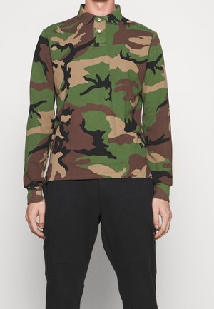 Personne portant un polo à manches longues à motif camouflage et un pantalon noir, debout devant un fond uni gris clair.