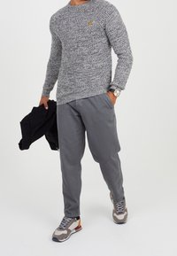 Szary sweter z dzianiny z małym żółtym logo, połączony z szarymi spodniami i wielokolorowymi trampkami. Czarna kurtka zarzucona na ramię.