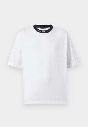 Młody mężczyzna stoi, ubrany w białą koszulę z krótkim rękawem z czarnym dekoltem, czarne szorty do kolan, białe skarpetki i czarne loafersy.