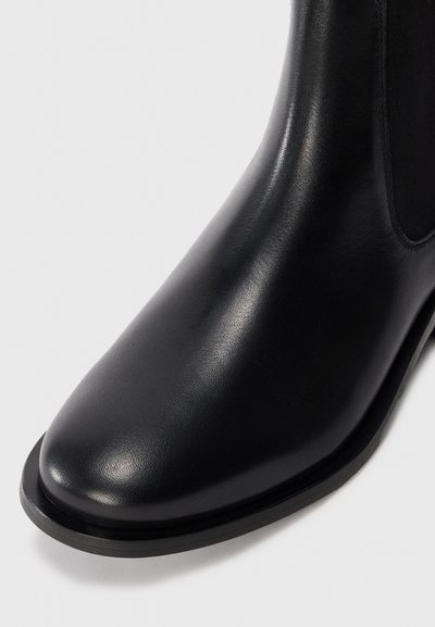 Svart läderankelboot med en slät yta, rund tå, elastiska sidopaneler och platt sula. Minimalistisk design utan synlig hårdvara.