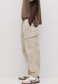 Beige cargobroek met elastische boorden, meerdere voorzakken en een trekkoord in de taille, gecombineerd met bruin-witte sneakers.