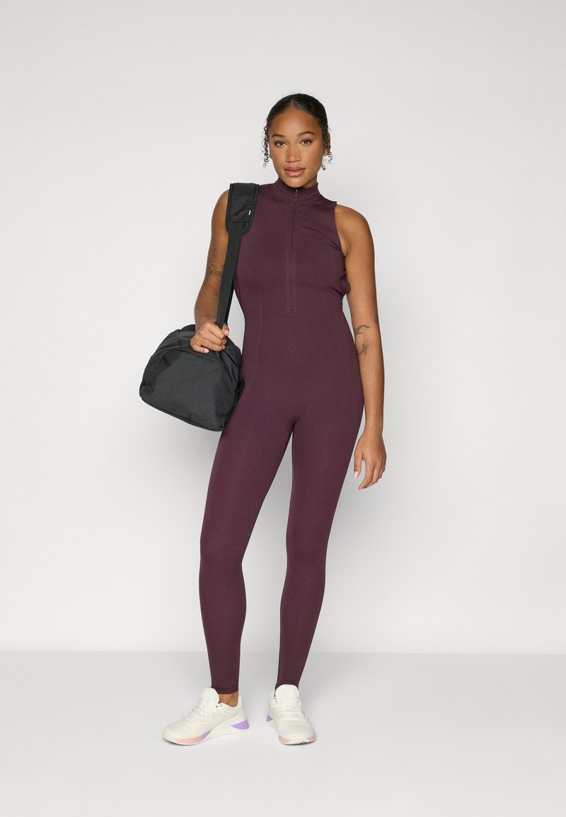 Maroon athletic jumpsuit with high collar and zipper, paired with a black duffel bag and white sneakers with purple accents.