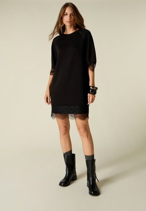Woman wearing black knee-length dress with lace trim, mid sleeves, black boots, gray socks, large black bracelets, and gold hoop earrings.