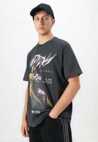 Young man wearing a black cap, dark Marvel graphic tee with Doctor Doom, and black Adidas pants with striped sides against a white background.