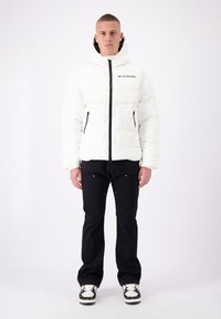 Witte pufferjack met capuchon, voorzien van een rits en zwarte accenten. Model draagt zwarte broek en sneakers, met een effen achtergrond.