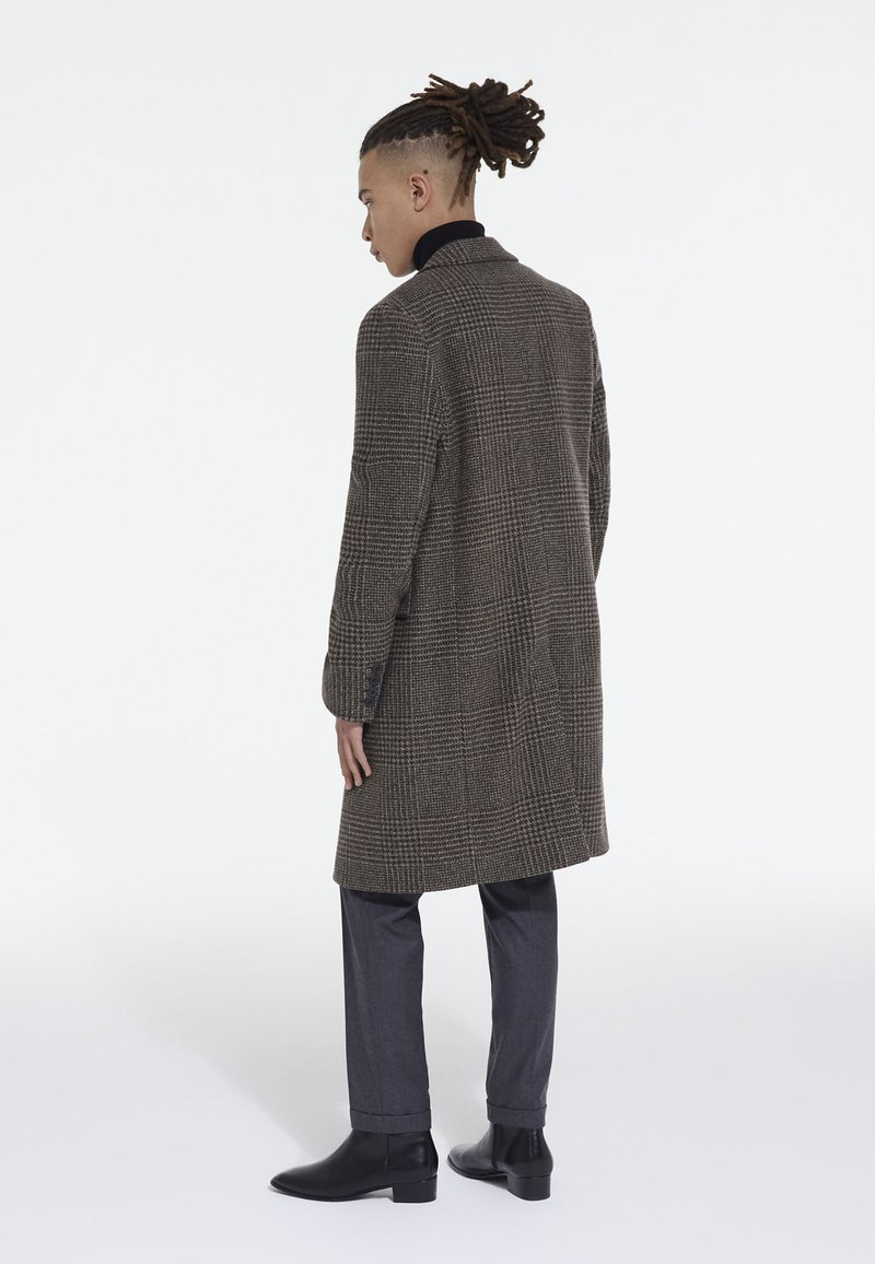 Cappotto lungo a quadri marrone in lana, abbinato a pantaloni grigi e stivali neri alla caviglia. Presenta spalle strutturate e una fila singola di bottoni.
