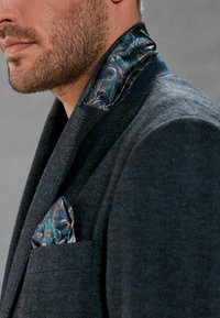 Blazer en chevrons foncé avec un col floral en satin et un pochettede couleur assortie. Tissu texturé avec des motifs complexes en bleu et vert.