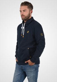Suéter de color azul marino con cuello alto, cordones blancos y detalles amarillos; cuenta con bolsillos frontales y un logo en el pecho y la manga.