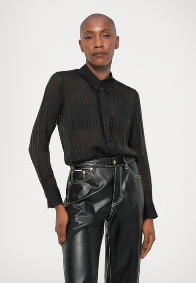 Schwarze durchsichtige Bluse mit vertikalen Streifen, einem Kragen und Knopfdetails, kombiniert mit hochgeschnittenen schwarzen Kunstlederhosen.